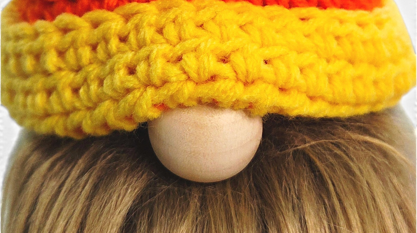 A close-up of a crocheted gnome hat with a textured orange top section, a wide yellow brim, and a light wood bead for a nose, shown on a person with blonde hair.