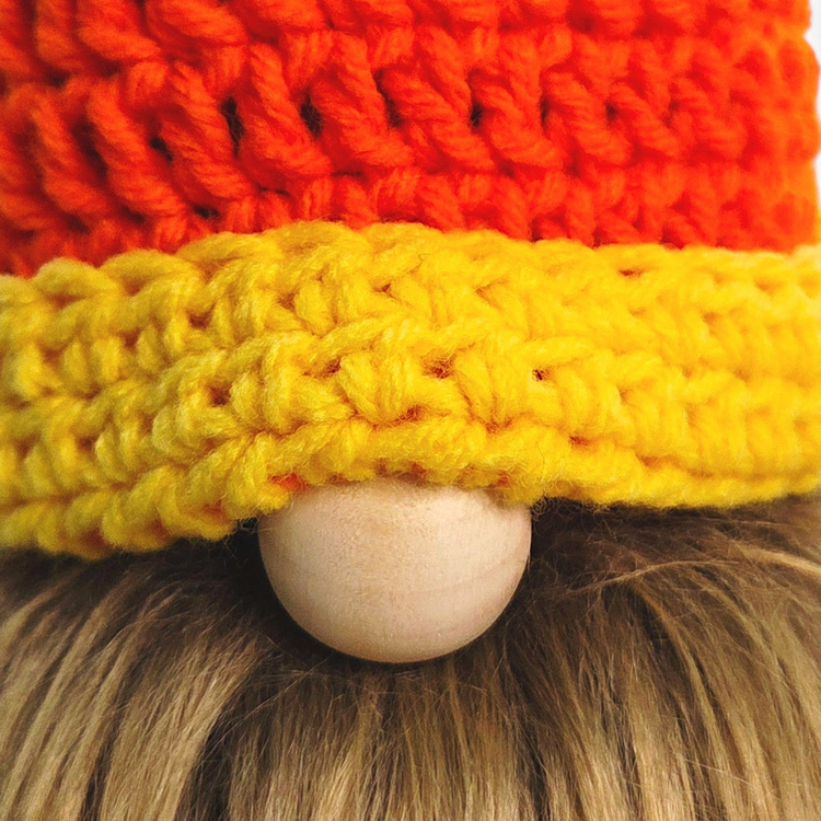 A close-up of a crocheted gnome hat with a textured orange top section, a wide yellow brim, and a light wood bead for a nose, shown on a person with blonde hair.