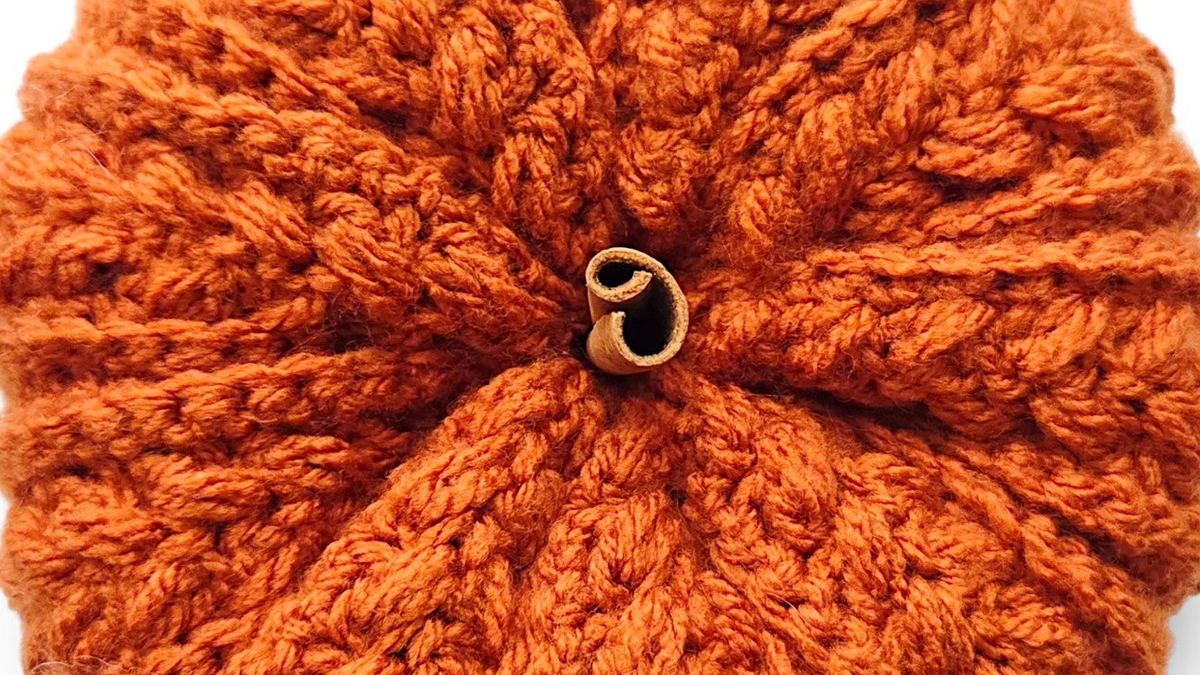 An orange crocheted pumpkin-shaped accessory with a small gold metal clasp at the top, shown on a clean white background.