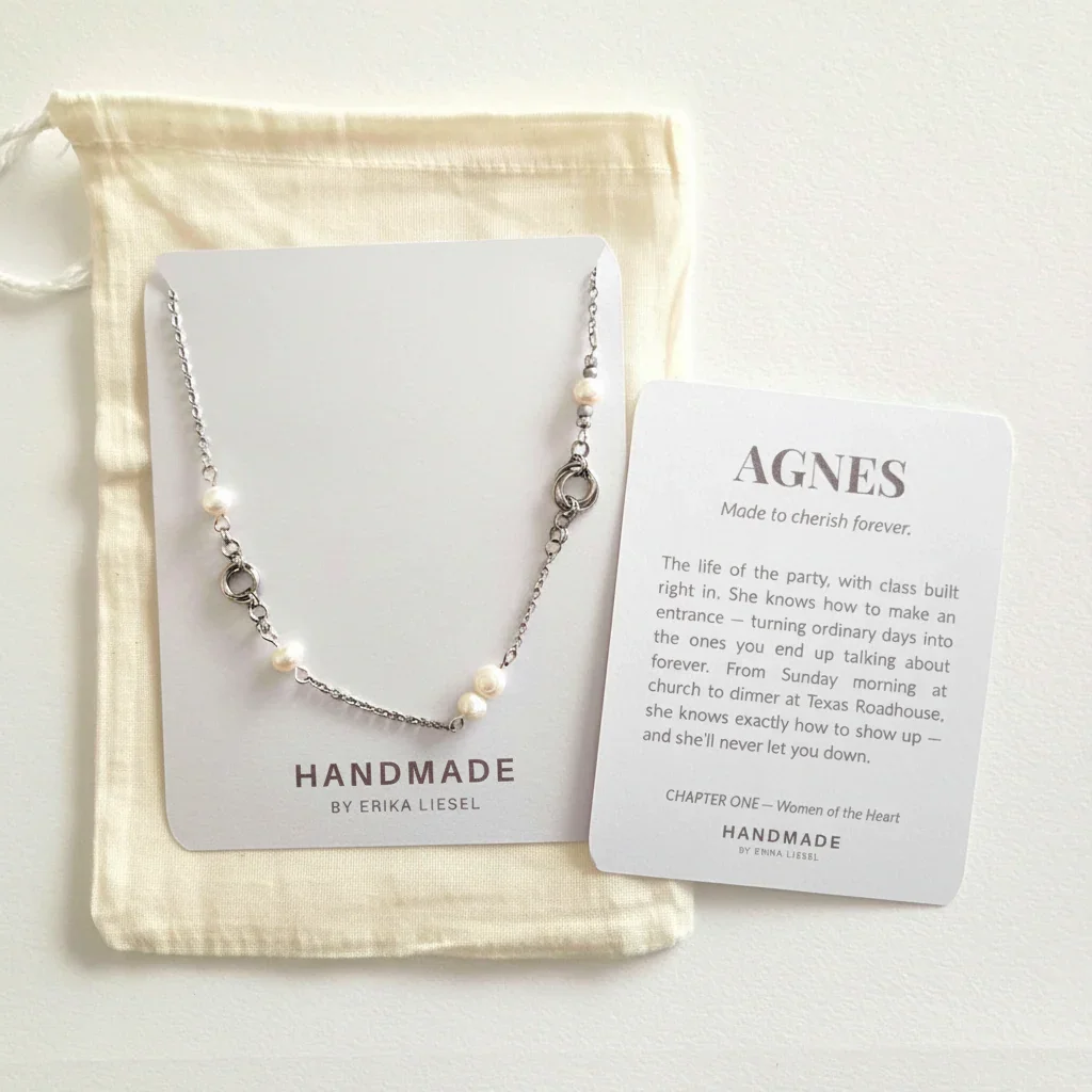 A delicate silver-toned necklace with small white pearl-like beads is displayed on a white card next to a cream drawstring pouch and a printed card reading 'AGNES' and 'HANDMADE BY ERIKA LIESEL'.