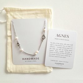 A delicate silver-toned necklace with small white pearl-like beads is displayed on a white card next to a cream drawstring pouch and a printed card reading 'AGNES' and 'HANDMADE BY ERIKA LIESEL'.