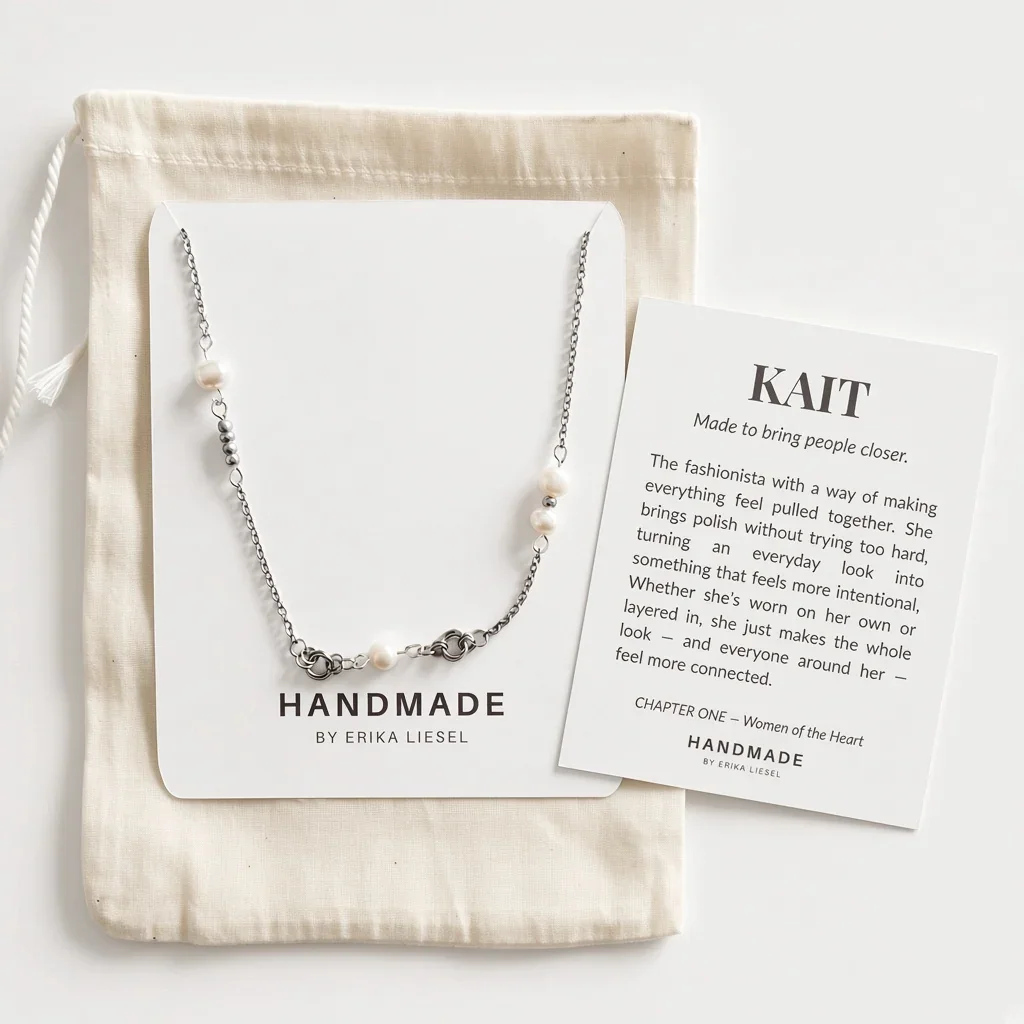 A silver beaded necklace with white pearl-like beads is displayed on a white card next to a cream drawstring pouch and a printed brand card. The necklace is shown in a styled flat-lay arrangement on a light neutral background.