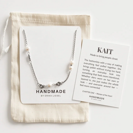 A silver beaded necklace with white pearl-like beads is displayed on a white card next to a cream drawstring pouch and a printed brand card. The necklace is shown in a styled flat-lay arrangement on a light neutral background.