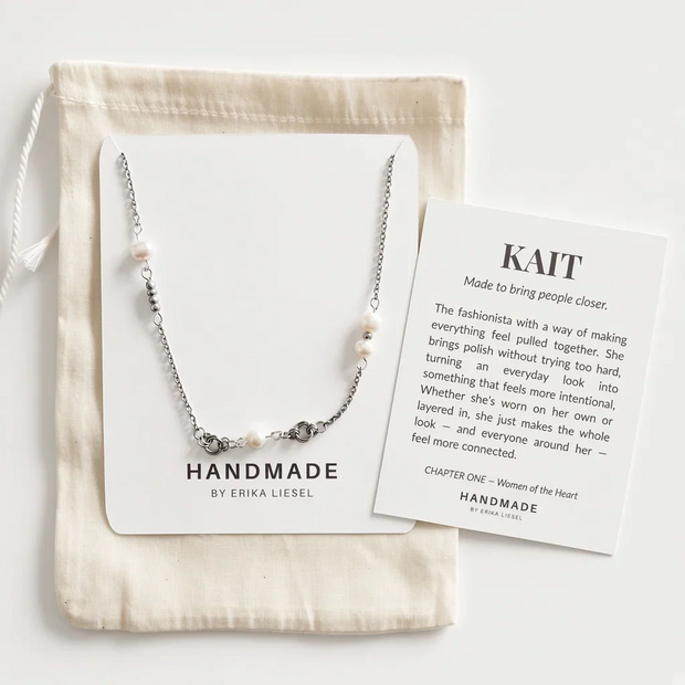 A silver beaded necklace with white pearl-like beads is displayed on a white card next to a cream drawstring pouch and a printed brand card. The necklace is shown in a styled flat-lay arrangement on a light neutral background.