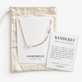 A delicate gold chain necklace with small clear beads, displayed on a white card next to a cream fabric drawstring bag and a printed gift message card. The brand HANDMADE BY ERIKA LIESEL is visible on the card.