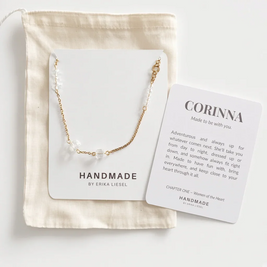 A delicate gold chain necklace with small white pearl-like beads displayed on a white card, placed on top of a cream fabric pouch. A second printed card reads “CORINNA” and includes a short message, photographed on a clean white background.