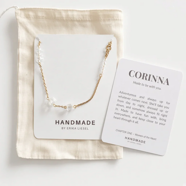 A delicate gold chain necklace with small white pearl-like beads displayed on a white card, placed on top of a cream fabric pouch. A second printed card reads “CORINNA” and includes a short message, photographed on a clean white background.