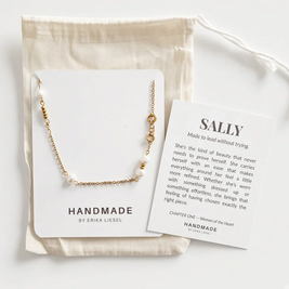 A delicate gold chain necklace with small white pearl-like beads and a gold oval charm, displayed on a white card next to a printed message card and a cream fabric pouch. The brand name 'HANDMADE BY ERIKA LIESEL' is visible on the packaging.