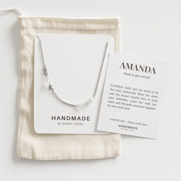 A delicate silver chain necklace with small clear beads and a central white pearl pendant, displayed on a white card next to a cream fabric pouch and a printed card reading 'AMANDA Made to get noticed.'.