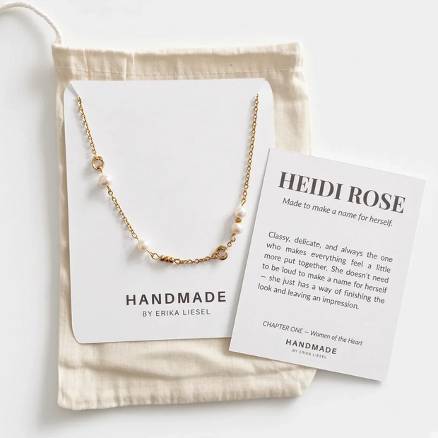 A delicate gold chain necklace with small white beads is displayed on a white jewelry card, next to a cream fabric pouch and a printed card titled 'HEIDI ROSE' with descriptive text. The brand 'HANDMADE BY ERIKA LIESEL' is visible on the jewelry card.