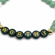 Handmade by Erika Liesel Worthy bracelet featuring green crystal beads and gold-plated letter beads on elastic cord.