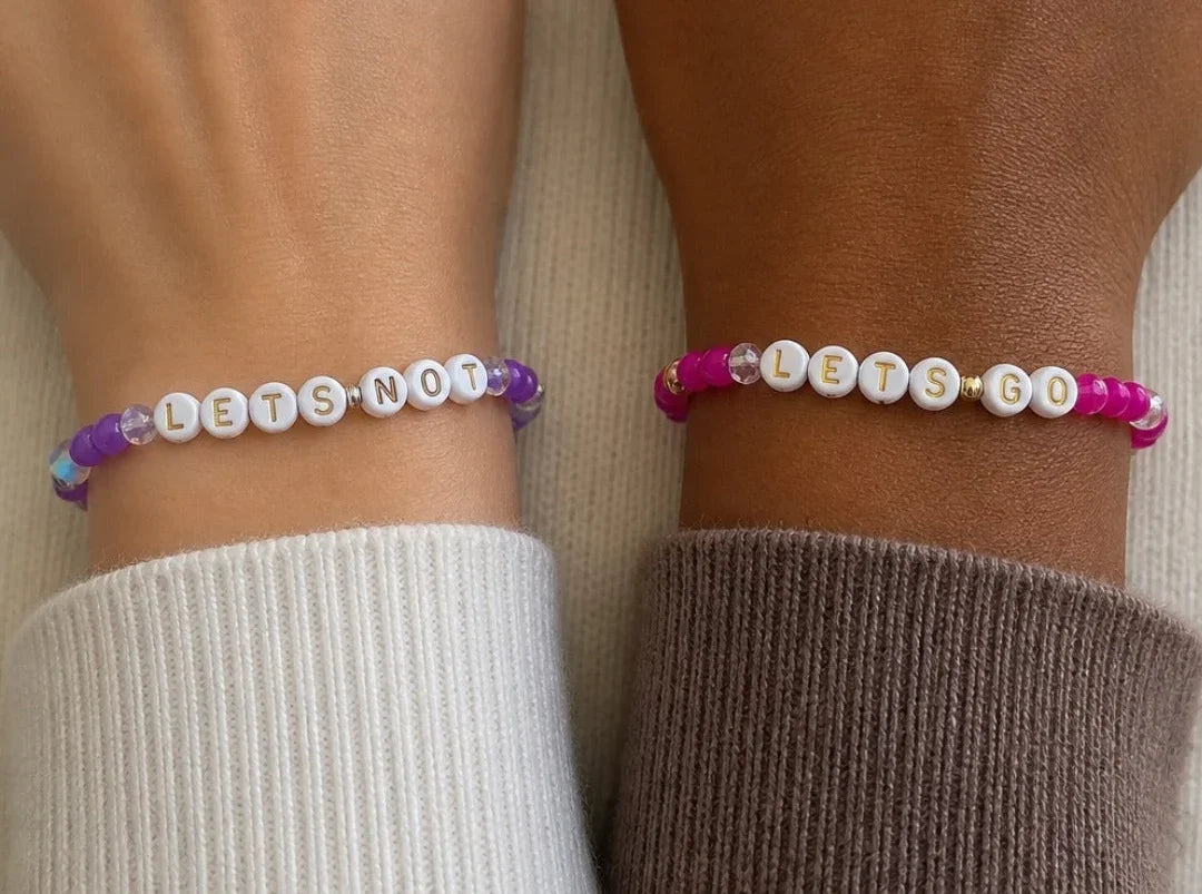 Two beaded bracelets worn on wrists, one with white beads spelling 'LET'S NOT' in purple and blue accents, and the other with white beads spelling 'LET'S GO' in pink and gold accents.