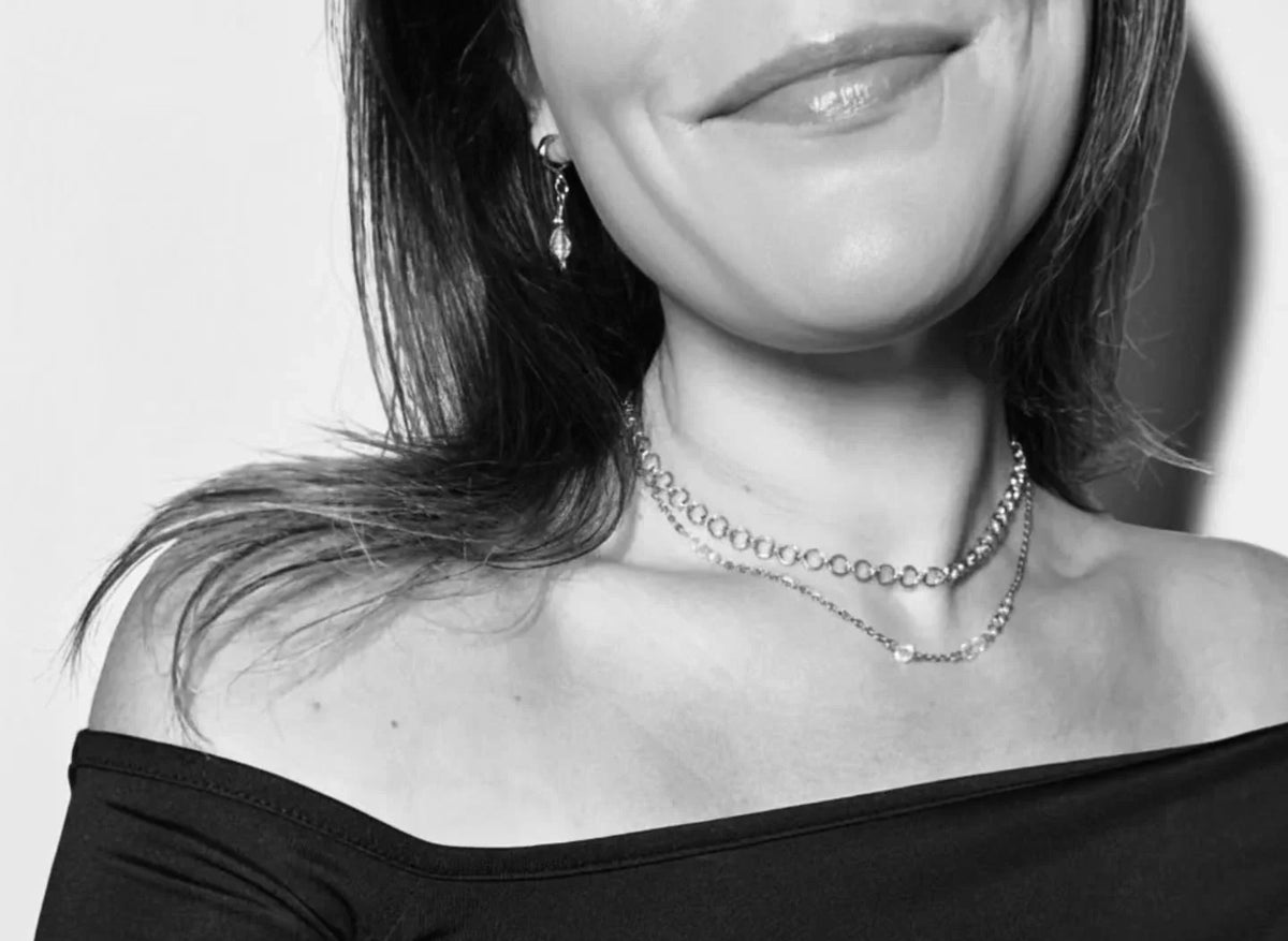A close-up black and white photo of a woman wearing a double-layered silver chain necklace and a matching drop earring.