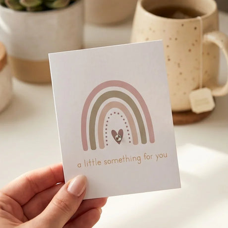 Handheld greeting card with pastel rainbow, heart, and 'a little something for you' text, cozy tea mug background