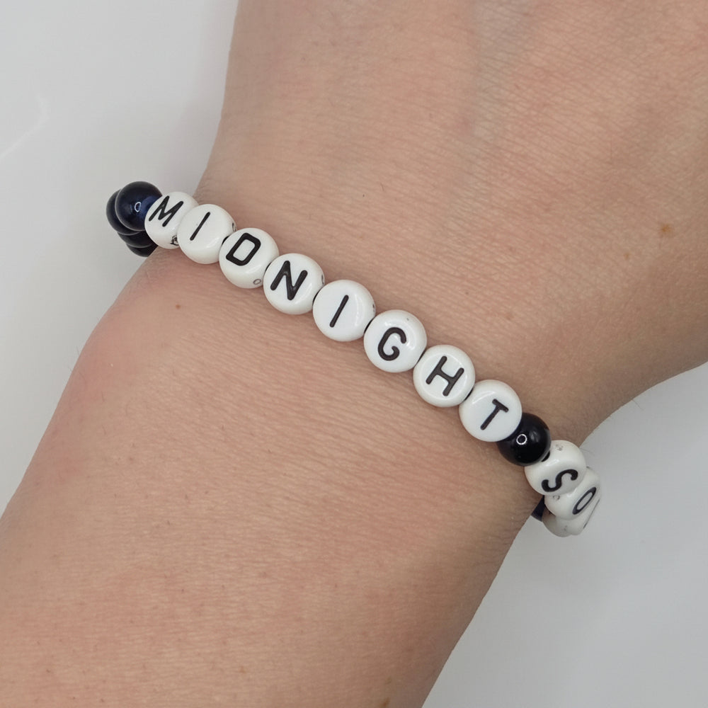 A beaded stretch bracelet worn on a wrist, featuring white letter beads spelling 'MIDNIGHT SO' and small black spacer beads, shown against a light neutral background.