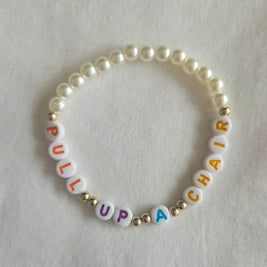 Pearl bracelet with colorful 'PULL UP A CHAIR' letter beads on white fabric