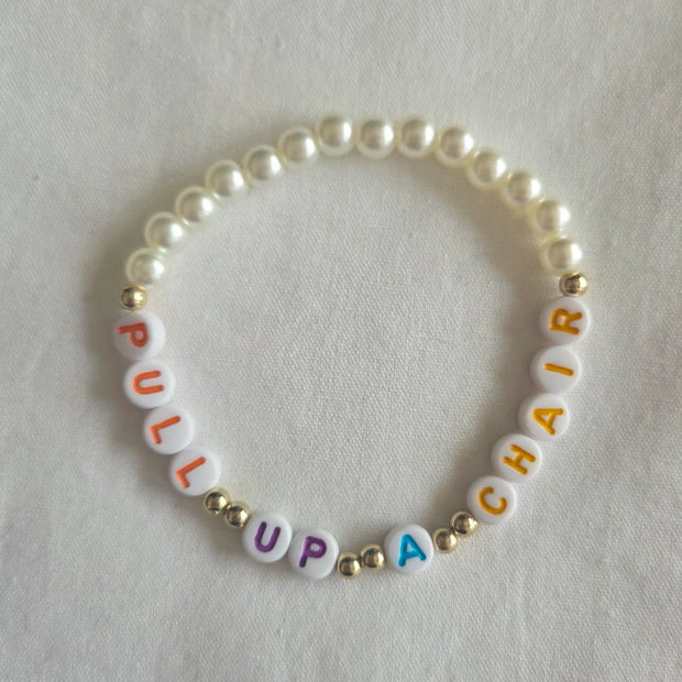 Pearl bracelet with colorful 'PULL UP A CHAIR' letter beads on white fabric