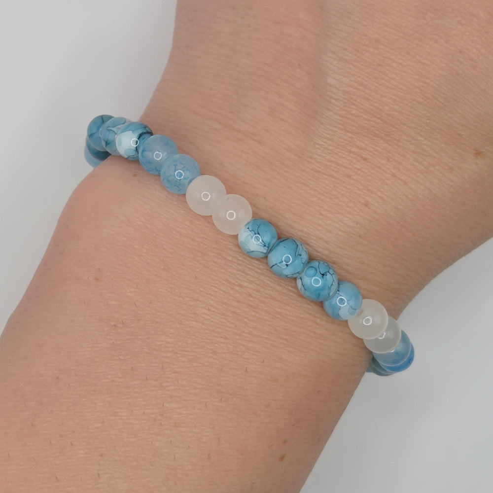 A beaded stretch bracelet worn on a wrist, featuring light blue marbled beads and translucent white beads, set against a plain light gray background.