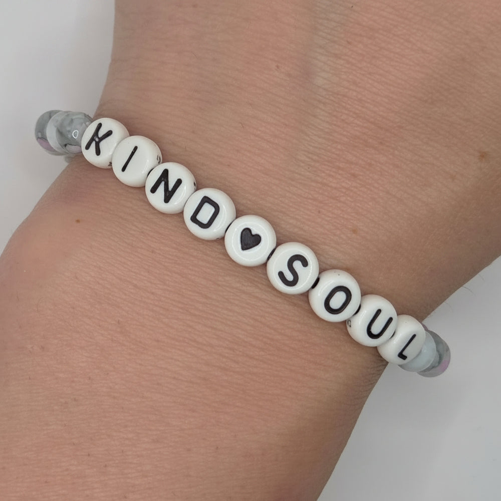 A beaded bracelet worn on a wrist, featuring white round letter beads spelling 'KIND SOUL' with a small black heart, and clear spacer beads on a light gray background.