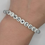 A beaded bracelet worn on a wrist, featuring white round letter beads spelling 'KIND SOUL' with a small black heart, and clear spacer beads on a light gray background.