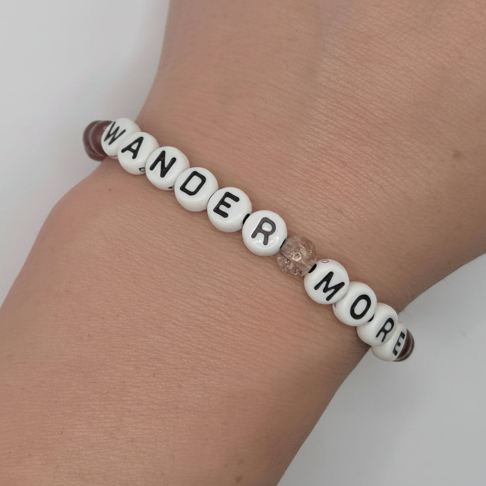 A beaded stretch bracelet worn on a wrist, featuring white round beads with black lettering that spell 'WANDER MORE', a small clear faceted bead, and dark brown end beads.