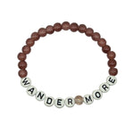 A beaded stretch bracelet featuring round, dusty rose-brown beads and white letter beads spelling out 'WANDER MORE' with a small textured beige accent bead in the center, displayed on a light gray background.