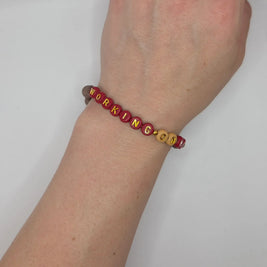 Red and gold beaded bracelet spelling 'WORKING ON IT' on wrist, handmade motivational accessory