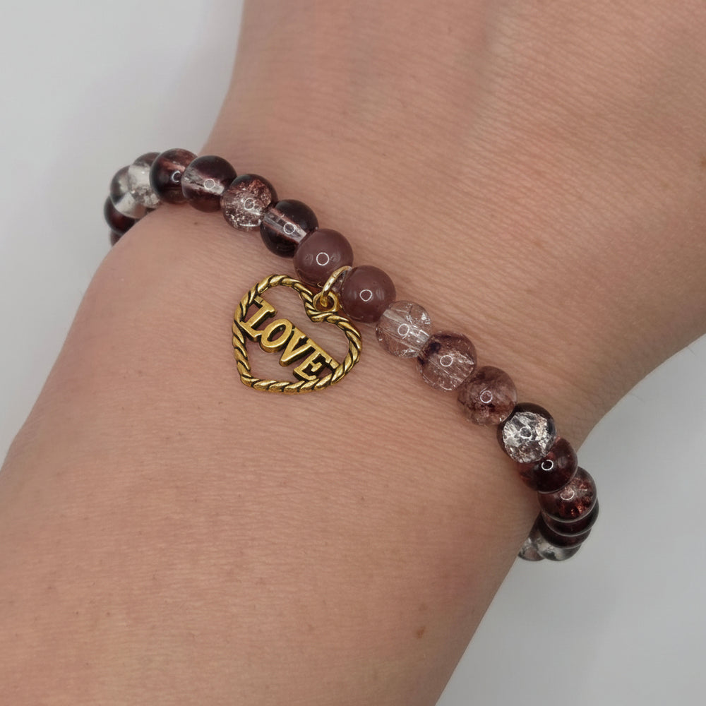 A close-up of a person's wrist wearing a beaded bracelet with round marbled dark purple and smoky gray beads, featuring a gold heart-shaped charm engraved with the word 'LOVE'.