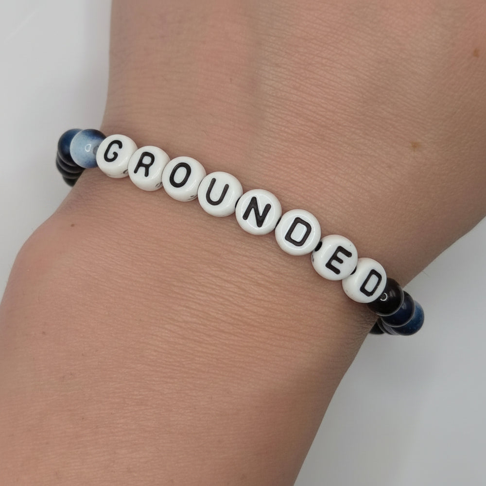 A close-up of a person's wrist wearing a beaded bracelet made of white letter beads spelling 'GROUNDED' with dark blue and black spacer beads on either end.