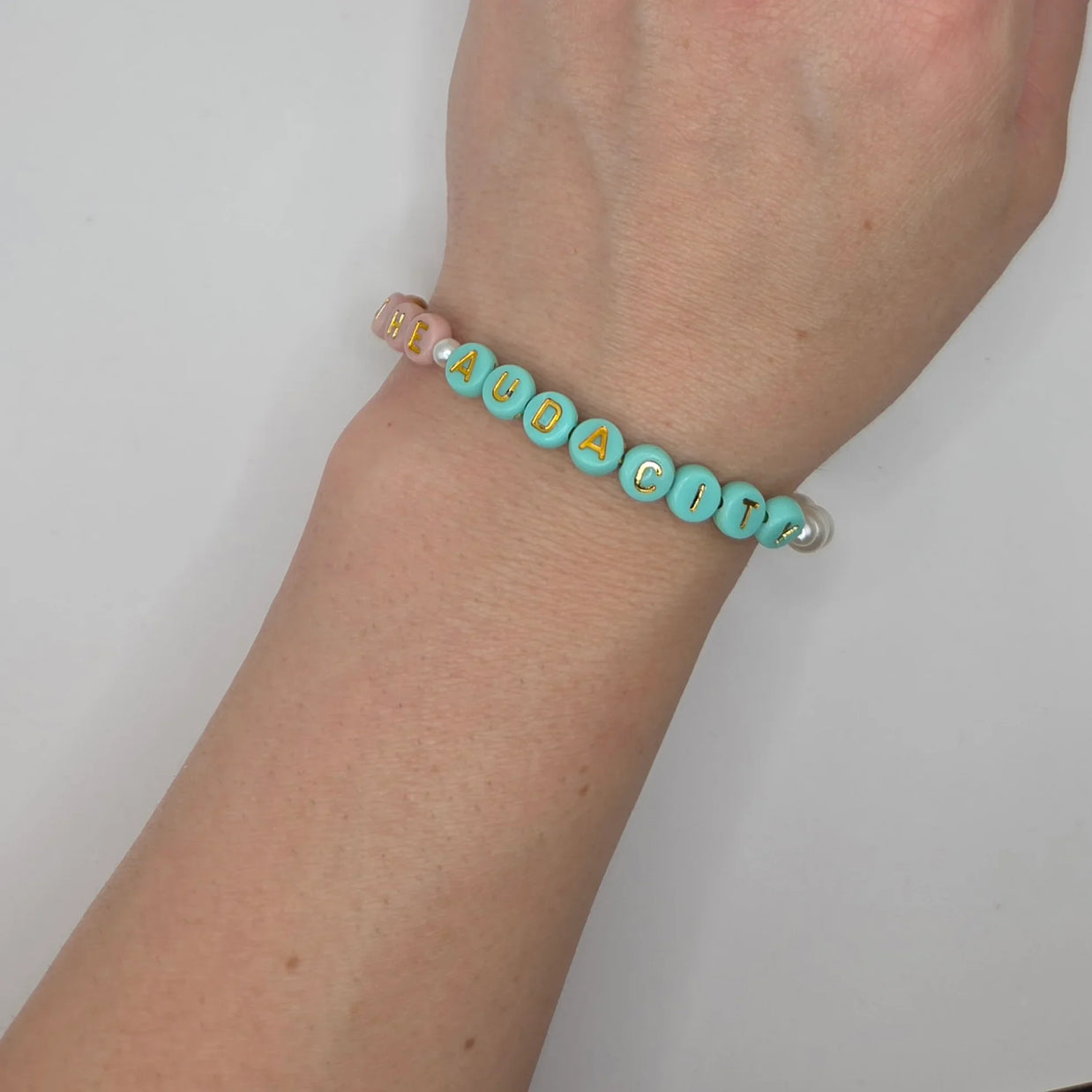Handmade beaded bracelet spelling 'HEAUDACITY' in gold letters on mint and pink beads, worn on wrist.