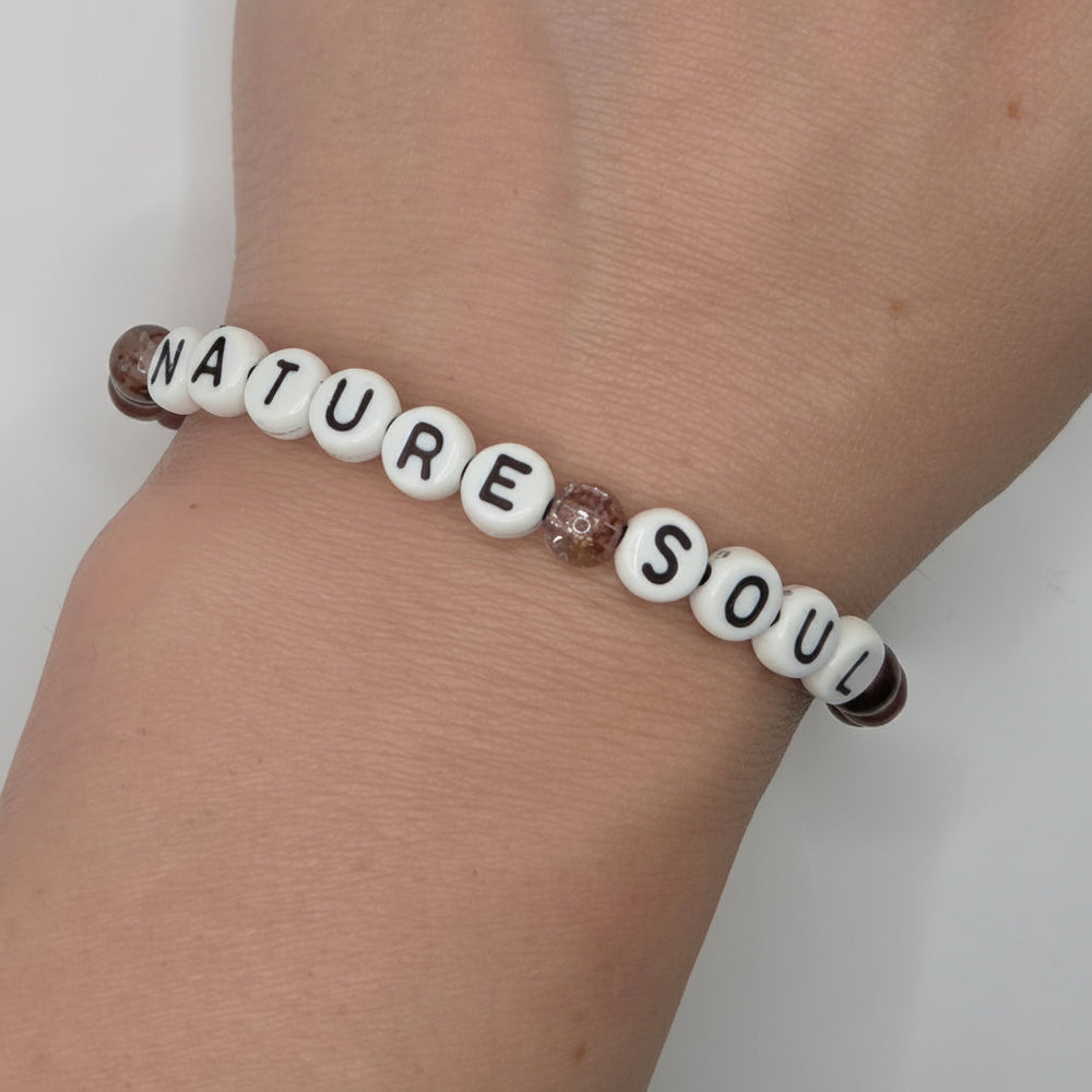 A beaded bracelet worn on a wrist, featuring white letter beads spelling 'NATURE SOUL' with brown textured beads on either end and a small glittery brown accent bead in the middle.