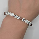 A beaded bracelet worn on a wrist, featuring white letter beads spelling 'NATURE SOUL' with brown textured beads on either end and a small glittery brown accent bead in the middle.