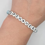A close-up of a person's wrist wearing a beaded bracelet made of white round letter beads spelling OPEN HEART, with a small light blue bead at one end.