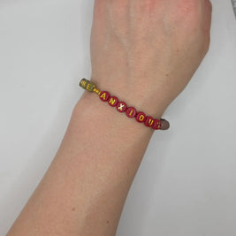 Handmade beaded bracelet spelling 'MEANXIOUS' in red and gold letters, worn on wrist for everyday style.