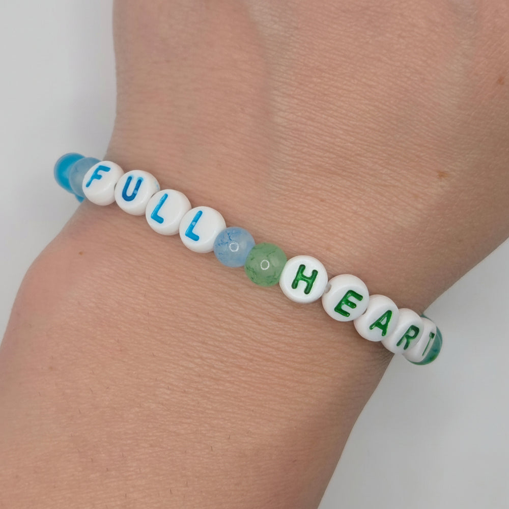 A beaded stretch bracelet worn on a wrist, featuring white letter beads spelling 'FULL HEART' with blue and green accents, and a translucent blue bead at one end.