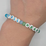 A beaded stretch bracelet worn on a wrist, featuring white letter beads spelling 'FULL HEART' with blue and green accents, and a translucent blue bead at one end.