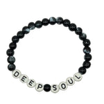 A beaded stretch bracelet on a plain light background. The bracelet features glossy black and white marbled beads, with white letter tiles spelling 'DEEP SOUL' at the front.