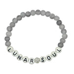 A beaded stretch bracelet on a white background, featuring gray marbled beads, white letter beads spelling 'LUNAR SOUL', and a small silver glitter accent bead.