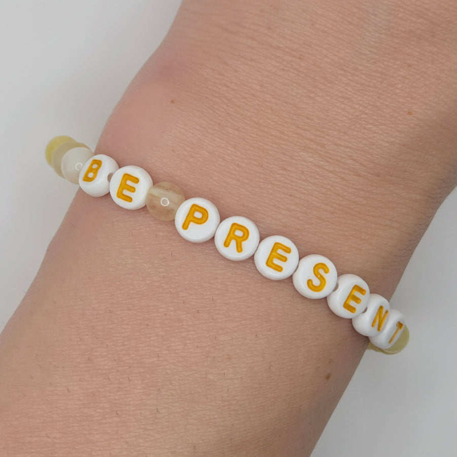A beaded stretch bracelet worn on a wrist, featuring white round beads with yellow letters spelling 'BE PRESENT' and a few translucent pale beads.