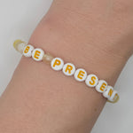A beaded stretch bracelet worn on a wrist, featuring white round beads with yellow letters spelling 'BE PRESENT' and a few translucent pale beads.