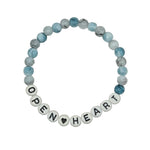 A beaded stretch bracelet on a light gray background. The bracelet features light blue and white marbled beads, with white letter beads spelling 'OPEN HEART' and one black heart bead.