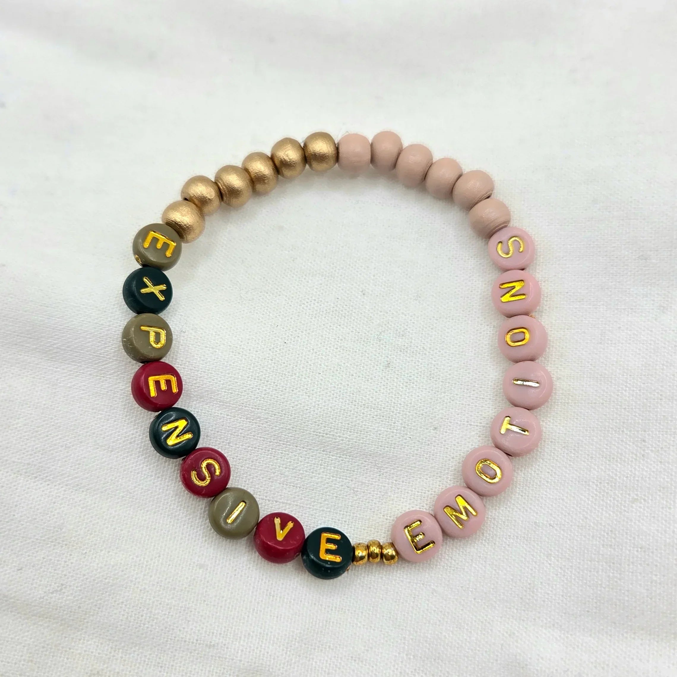 Handmade beaded bracelet with 'EXPENSIVE EMOTIONS' in colorful letters, gold accents, soft pink beads