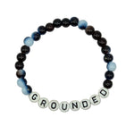 A beaded stretch bracelet on a light gray background, featuring dark brown and black beads, light blue and white marbled beads, and white letter beads spelling 'GROUNDED'.