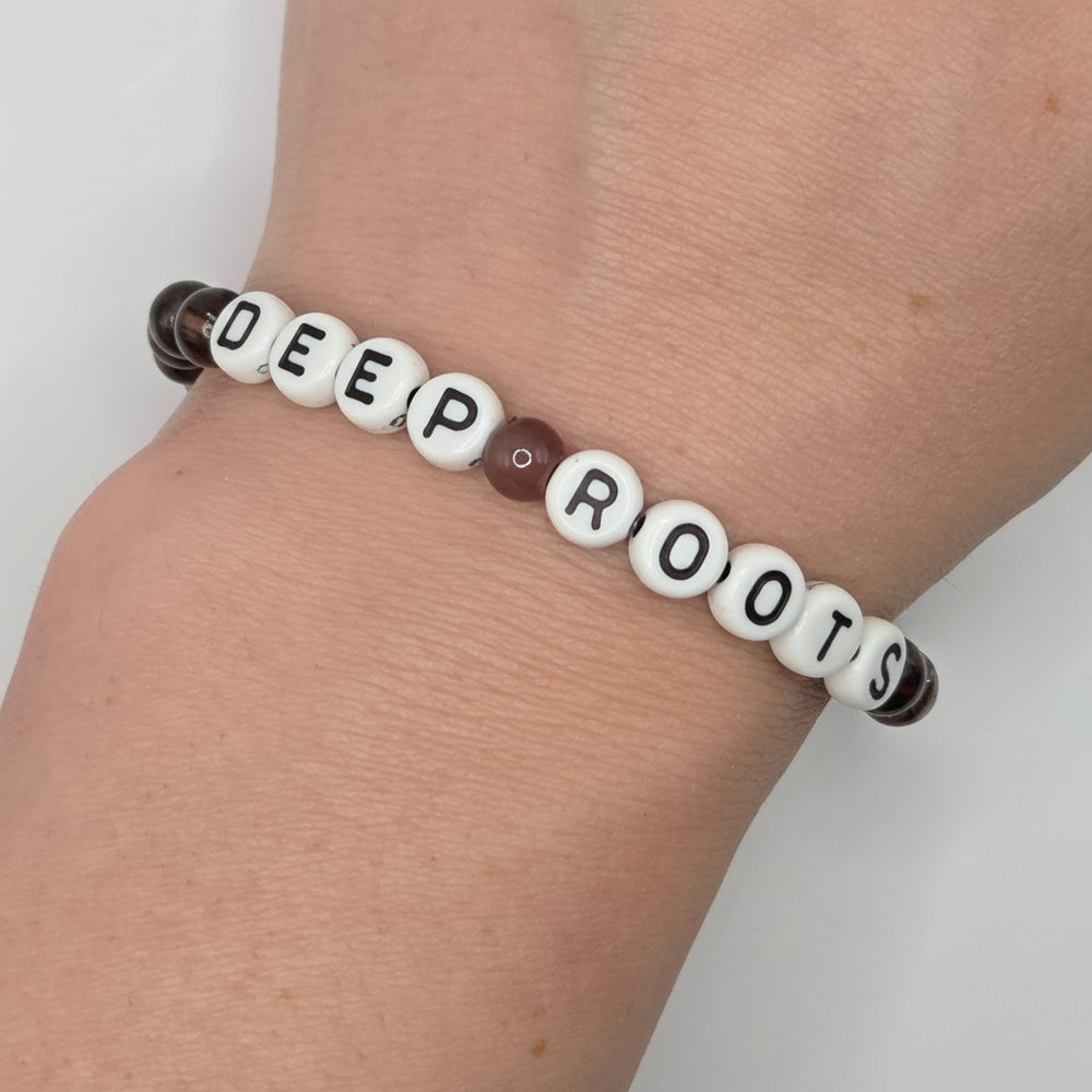 A beaded stretch bracelet spelling 'DEEP ROOTS' worn on a person's wrist. The beads are primarily white with black lettering, one brown bead, and two dark smoky marbled end beads.