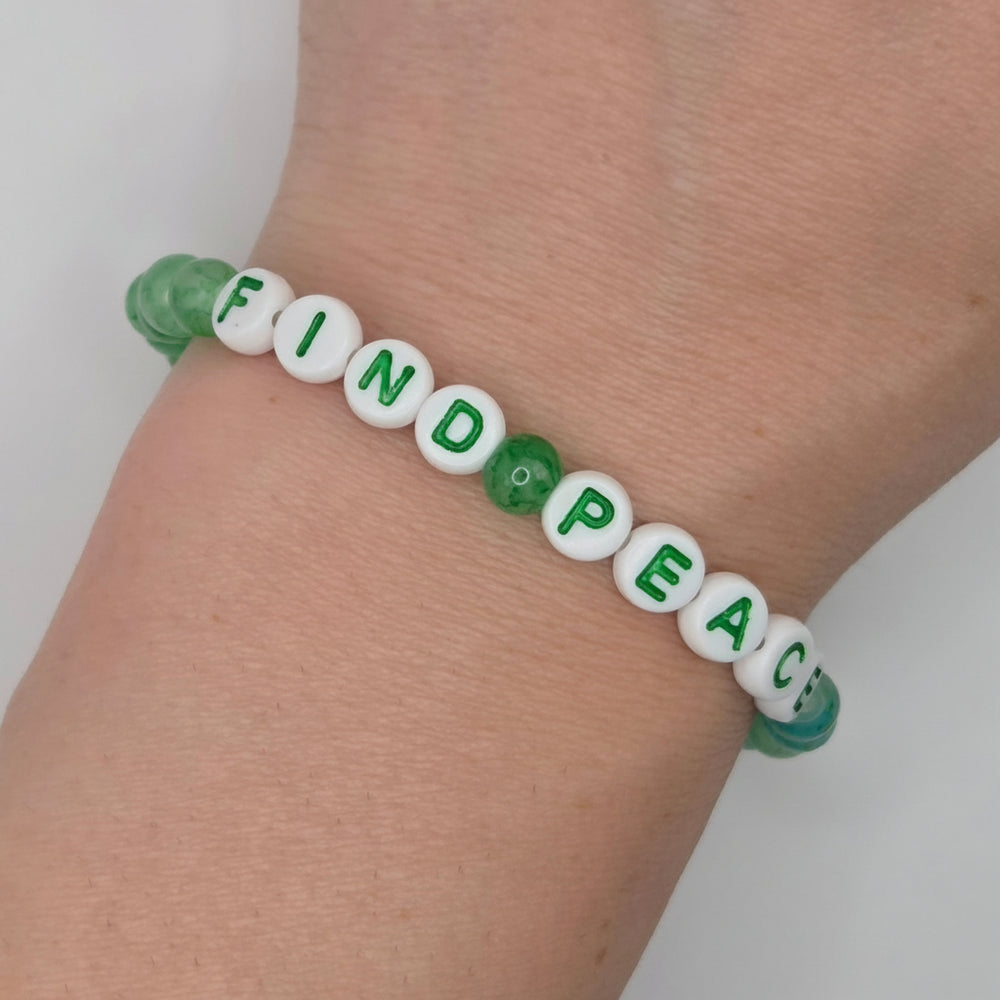 A close-up of a person's wrist wearing a beaded bracelet made of white letter beads spelling 'FIND PEACE' with green lettering, plus green translucent beads and a green flower-shaped bead.