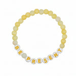 A beaded stretch bracelet on a plain white background, featuring pale yellow and off-white round beads with white letter beads spelling 'BE PRESENT' in yellow.