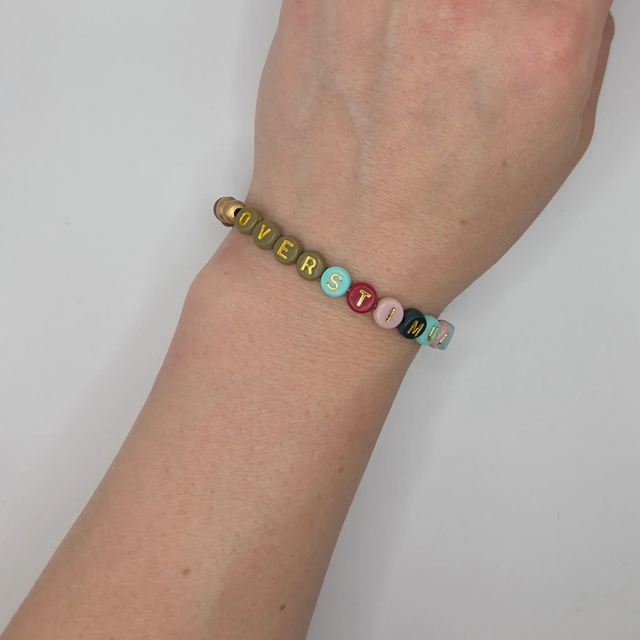 Colorful beaded bracelet spelling 'OVERSTIMULATED' on wrist, handmade gift for cozy comfort