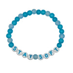 A beaded stretch bracelet arranged in a circle on a light gray background. The bracelet features alternating translucent light blue and opaque medium blue beads, with white letter beads spelling out 'STAY SOFT' in blue lettering.