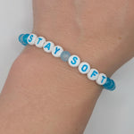 A close-up of a person's wrist wearing a beaded stretch bracelet made of white round beads with blue lettering spelling 'STAY SOFT', plus translucent blue beads at the ends.