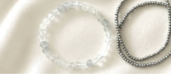 Two beaded stretch bracelets displayed on a soft white fabric background. One bracelet is made of translucent white round beads, and the other is made of small metallic silver beads.